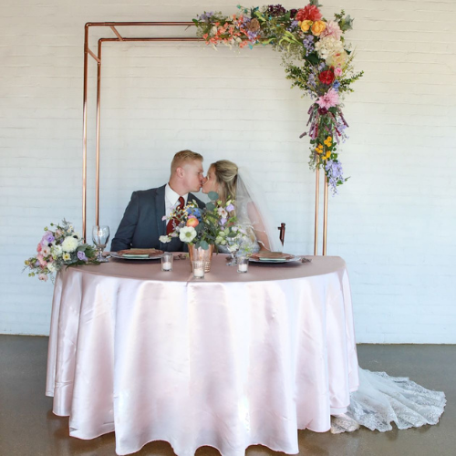 Bride and groom kissing while sat at a set table in front of a wedding arch